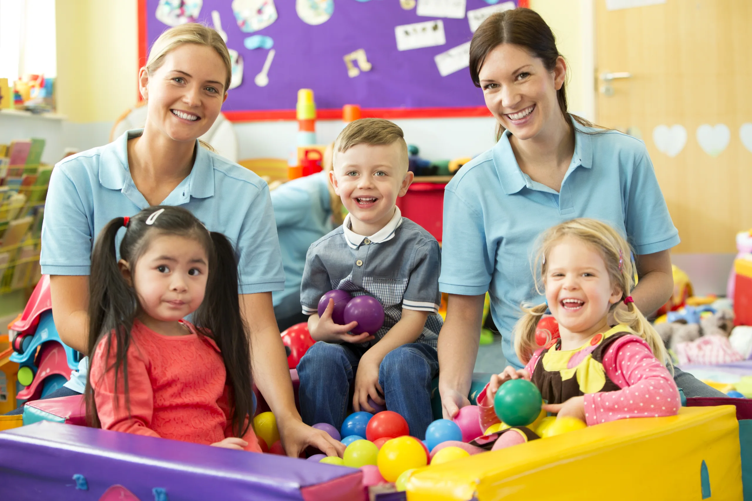 A horizontal image of three children in a ball pool playing and laughing, behind them sit two nursery teachers in blue polo shirts supervising and smiling. The nursery is a colourful scene of toys and mess.